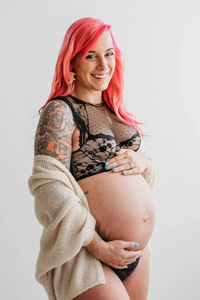 a photo of a pregnant woman with pink hair wearing black lingerie and looking at the camera and smiling, posing for a professional boudoir maternity shoot at a pamela erwin photo studio in Glasgow