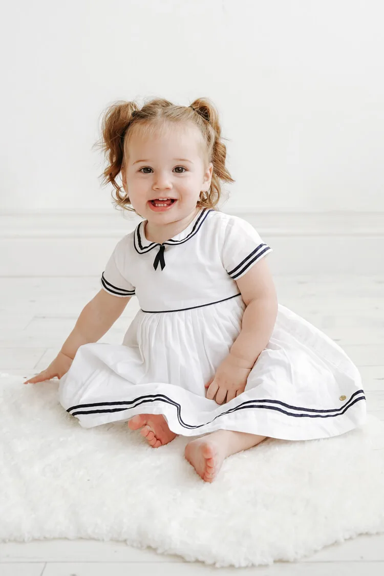 professional photo of a two year old little girl wearing a white dress sitting on the white floor, smiling and looking into the camera at a professional photography studio in glasgow